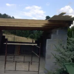 Budowa zadaszenia z bloczków betonowych i drewnianej konstrukcji dachowej, widoczne rusztowanie i drzewa w tle.