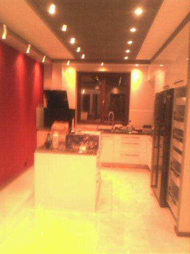 Nowoczesna kuchnia z wyspą, oświetleniem punktowym i czerwoną ścianą akcentującą, widziana z perspektywy wejścia.