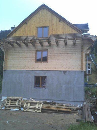 Surowy stan domu murowanego z drewnianą elewacją w trakcie budowy, widoczne drewniane elementy konstrukcyjne, szara dolna część ściany, palety i deski leżące na ziemi.