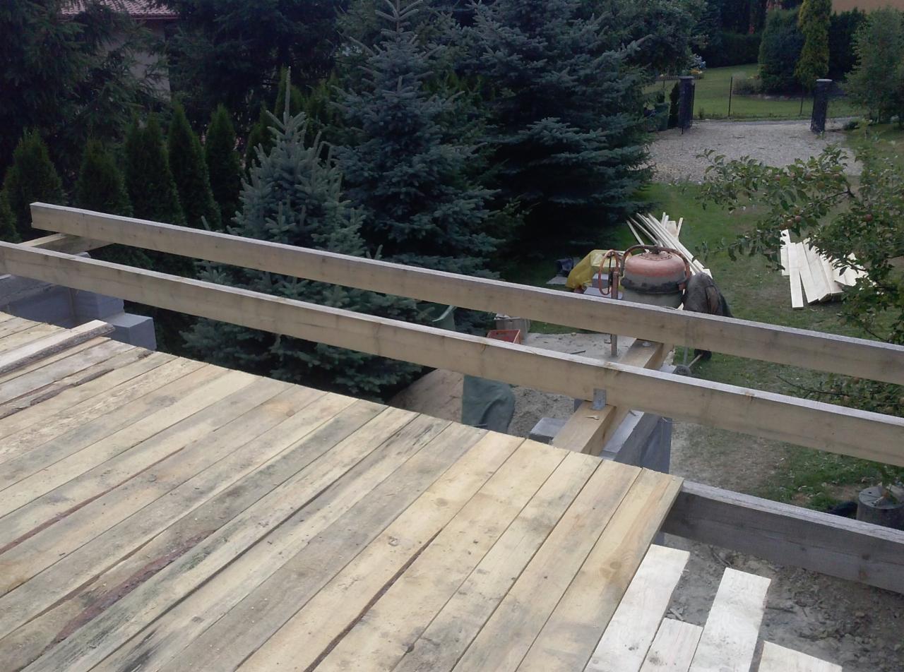 Wiązanie drewnianych belek na budowie domu jednorodzinnego, widoczna betoniarka i składowane deski na tle zieleni.