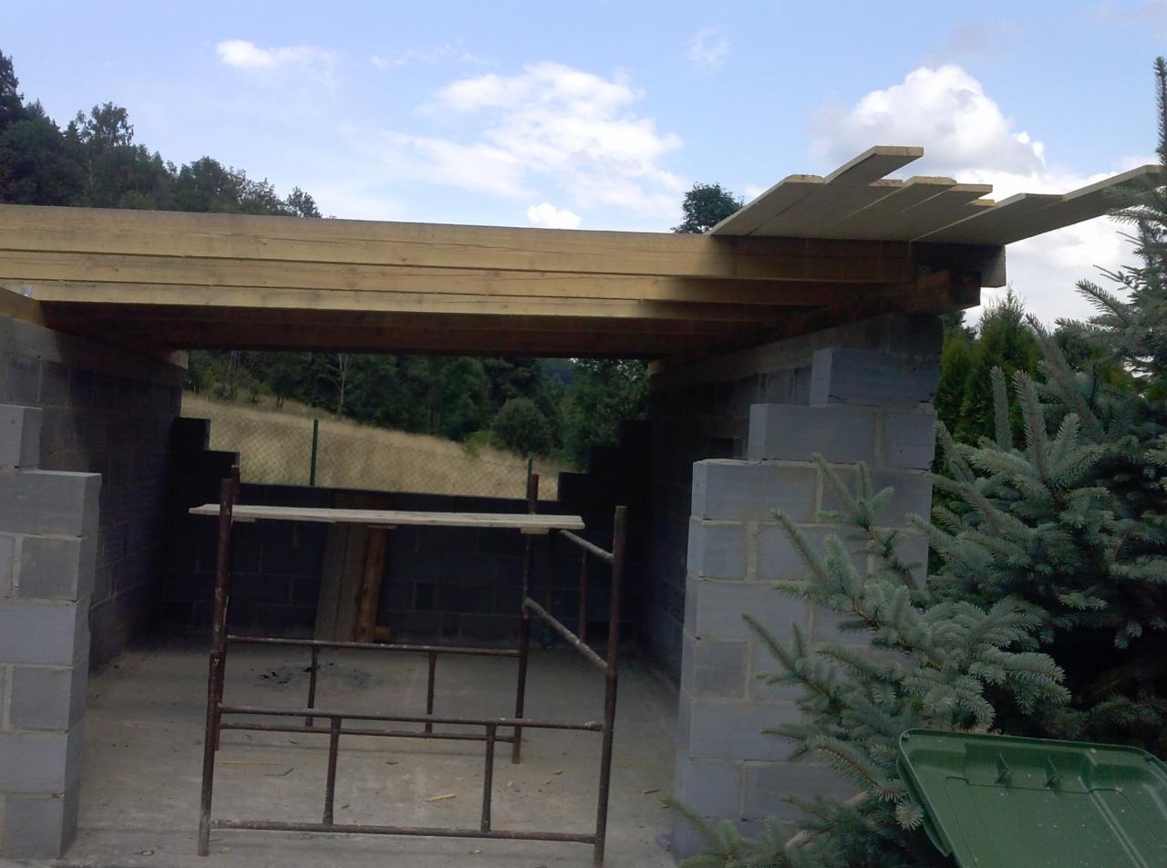 Budowa zadaszenia z bloczków betonowych i drewnianej konstrukcji dachowej, widoczne rusztowanie i drzewa w tle.