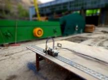 Precyzyjny wózek pomiarowy z czujnikami na szynie pomiarowej, ustawiony na drewnianej płycie, w tle zielona konstrukcja stalowa i fragment budynku.