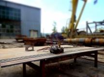 Metalowe elementy i narzędzia na drewnianym stole warsztatowym na zewnątrz, w tle budynek przemysłowy i fragment żurawia.