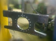 Metalowy element w kształcie litery H z wyciętym okręgiem i otworami, zawieszony na lince, z rozmytym tłem warsztatowym.