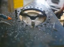 Metalowa płyta z wyfrezowanym okrągłym otworem i licznymi mniejszymi otworami wokół, widoczne ślady obróbki i chłodziwo.