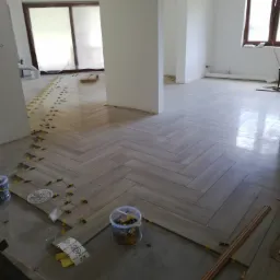 Układanie podłogi z paneli w jodełkę, widoczne plastikowe kliny dystansowe i materiały budowlane, surowy stan wykończenia ścian i sufitu, duże okna.