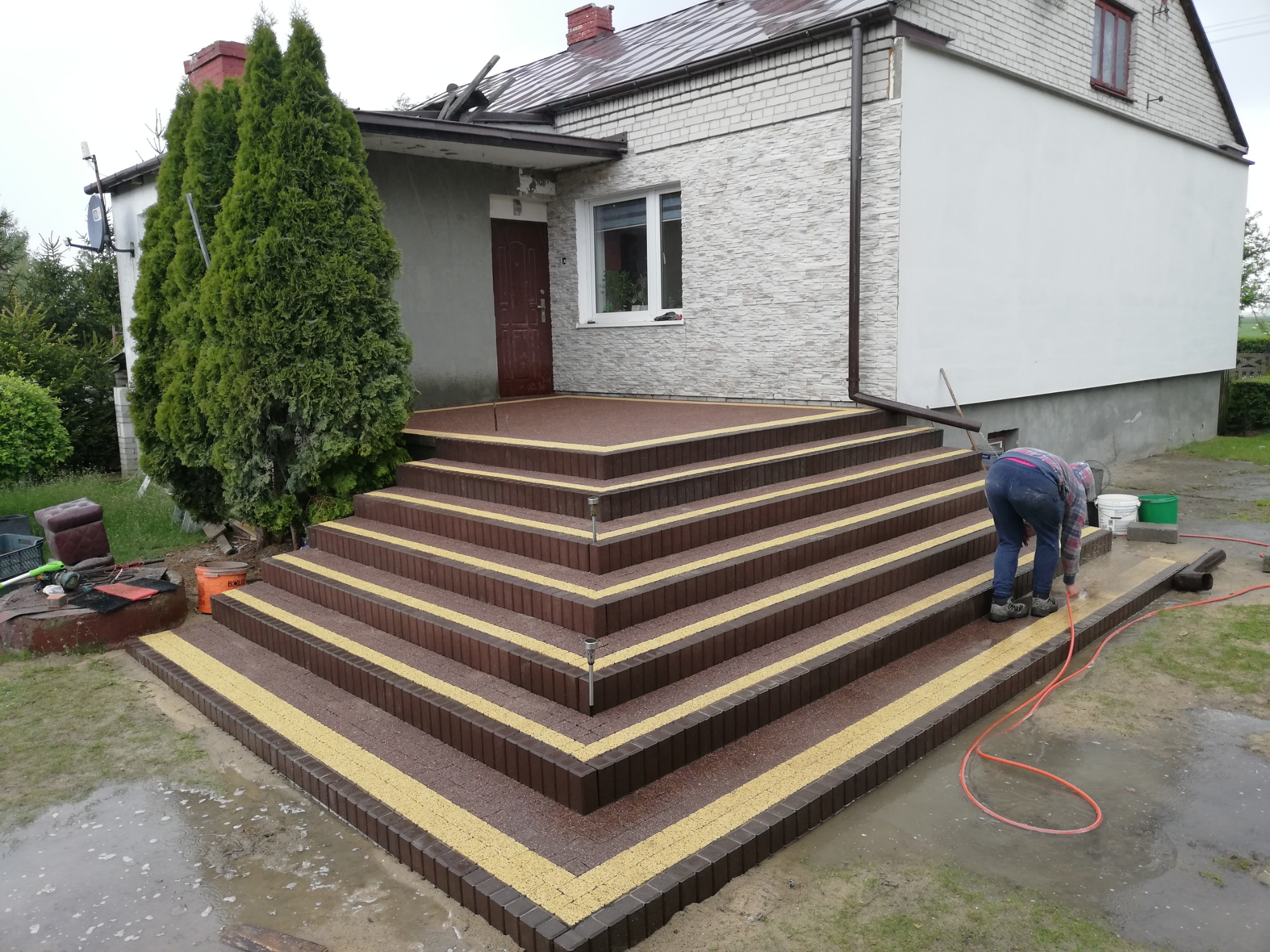 Montaż schodów zewnętrznych z kostki brukowej w kolorze brązowo-żółtym, pracownik w trakcie czyszczenia powierzchni, dom jednorodzinny w tle, deszczowa pogoda.