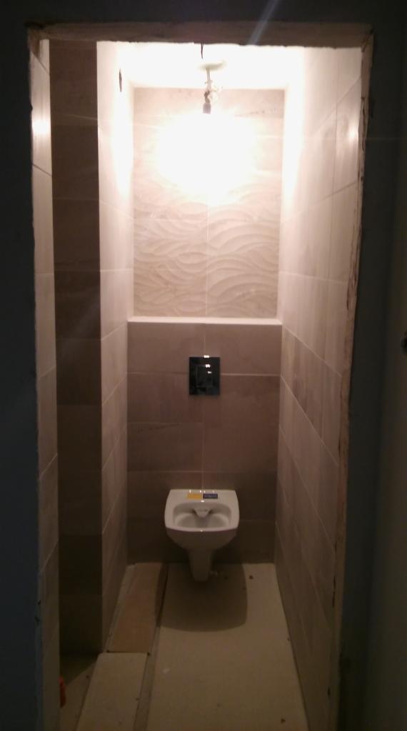 Wąska toaleta w trakcie wykańczania, widoczne płytki w odcieniach beżu, podwieszana muszla klozetowa z żółtą naklejką, surowa ściana przy wejściu i prowizoryczne oświetlenie.