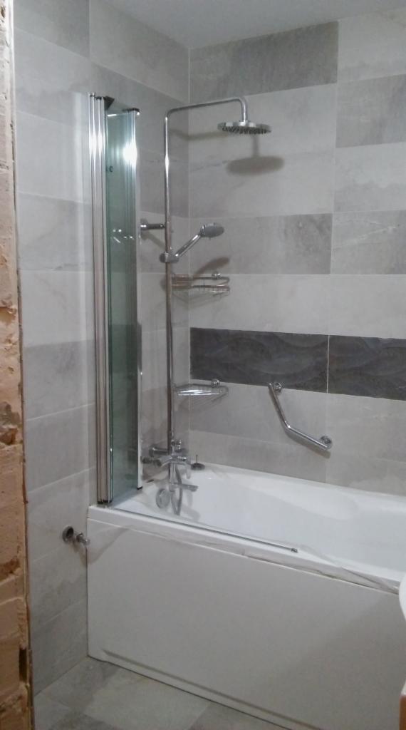 Wyremontowana łazienka z białą wanną, szarą glazurą w poziome pasy z ciemniejszym akcentem, szklanym parawanem nawannowym i chromowaną armaturą, widoczny fragment surowej ściany po lewej stronie.