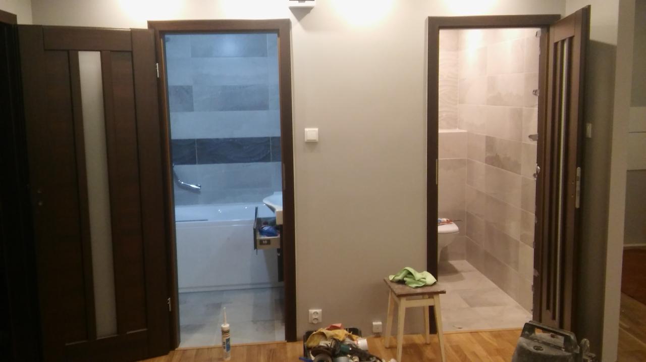 Dwa otwarte, drewniane drzwi prowadzące do wykafelkowanej łazienki i pokoju z wanną, widoczne narzędzia i materiały na podłodze, krzesło z zieloną szmatką.