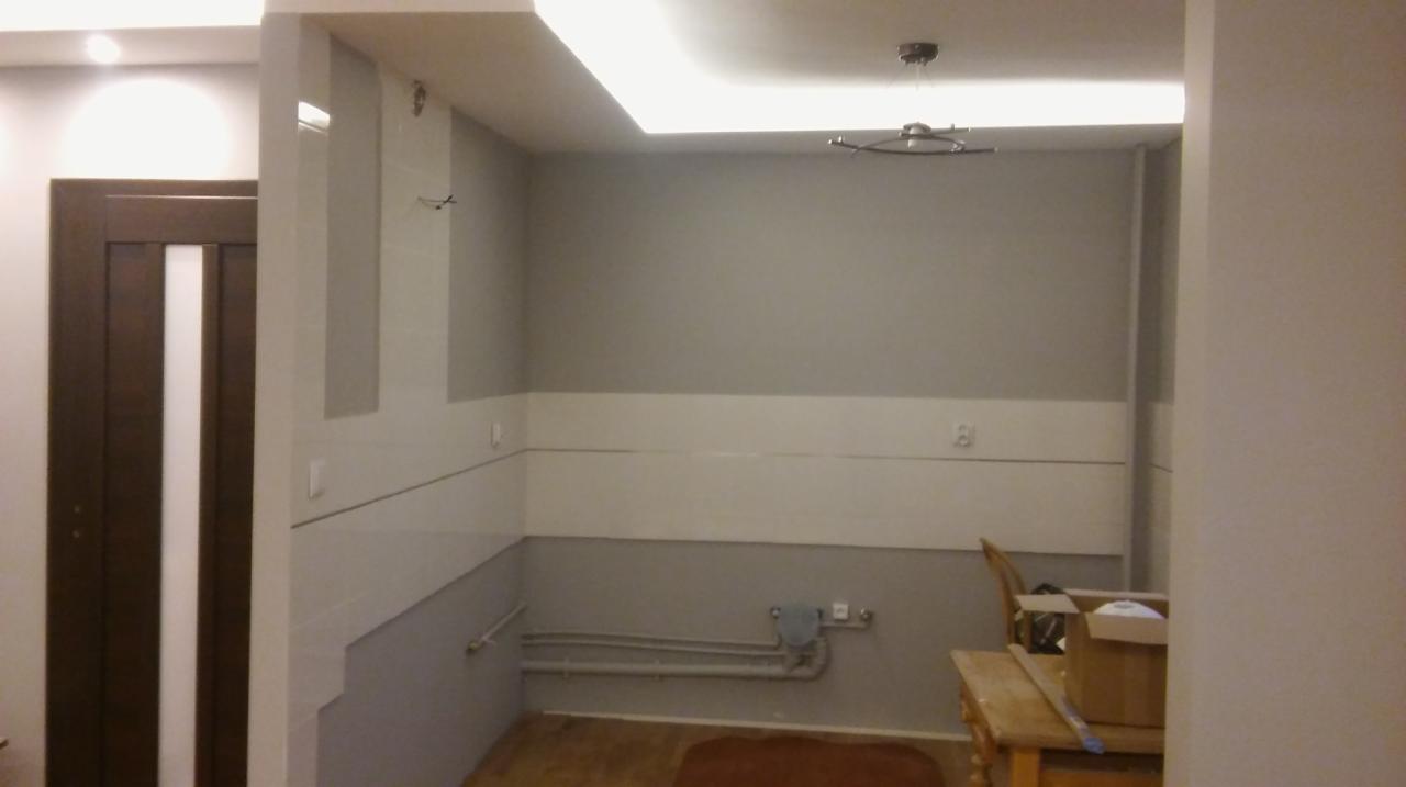 Wnętrze pomieszczenia w trakcie prac: szare ściany z poziomym pasem białych płytek, widoczne rury przy podłodze, drewniane drzwi z białym pasem, sufit z oświetleniem ledowym.