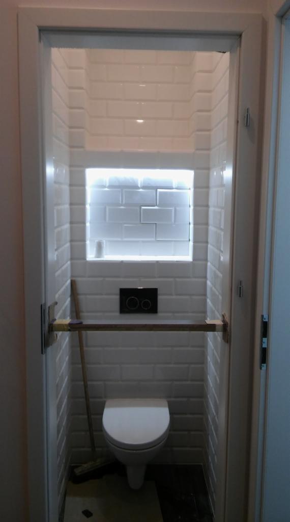 Wąska toaleta z białymi, cegiełkowymi płytkami, podświetloną wnęką i deską opartą o ściany, widok z zewnątrz.