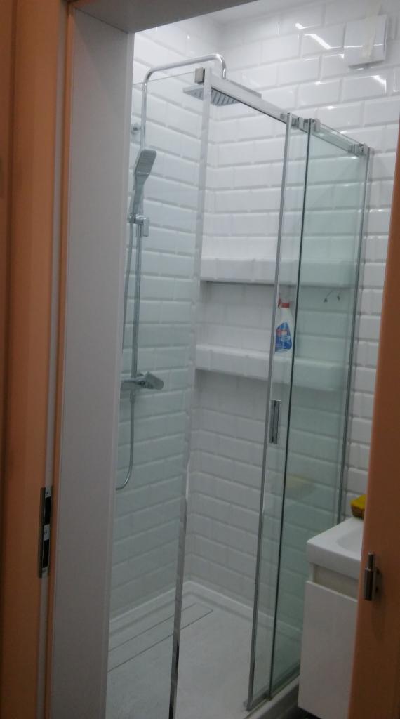 Nowoczesna kabina prysznicowa z przesuwnymi szklanymi drzwiami, wyłożona białymi, prostokątnymi płytkami imitującymi cegłę, z widoczną półką z kosmetykami.