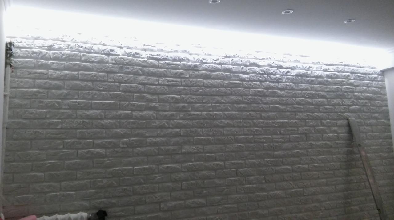 Ściana wykończona szarą, dekoracyjną cegłą z widocznym oświetleniem LED pod sufitem i fragmentem drabiny opartą o ścianę.