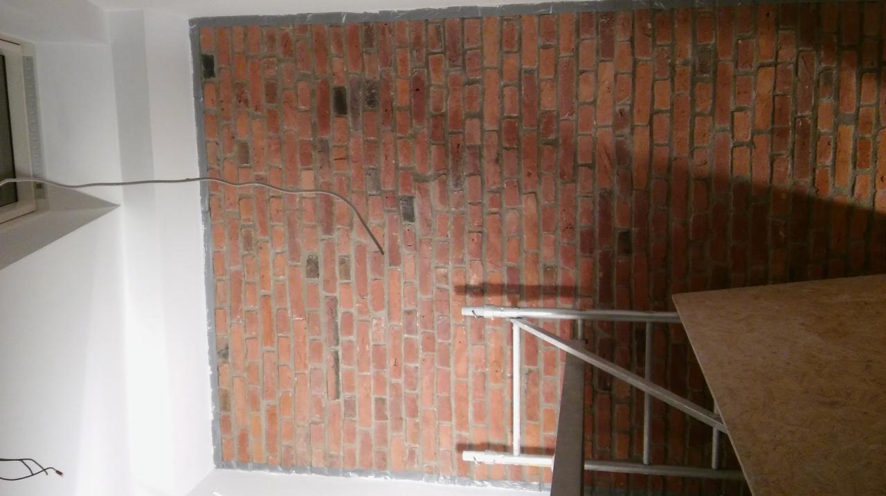 Ściana z czerwonej cegły w trakcie wykańczania, widoczne przewody elektryczne i fragment rusztowania.