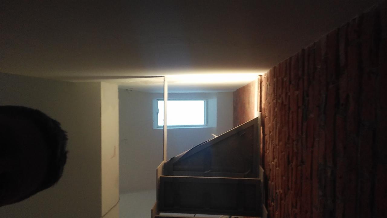 Wnętrze w trakcie prac: surowa ściana z cegły, widoczne schody w drewnianej obudowie oraz okno w tle, częściowo zakryte przez pionowy element konstrukcyjny.