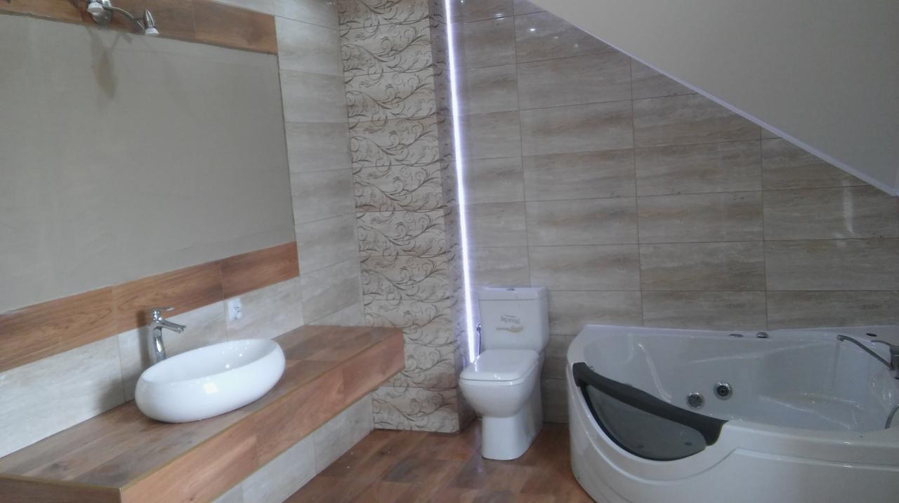 Jasna łazienka z wanną z hydromasażem, umywalką nablatową na drewnianym blacie i toaletą; ściany wyłożone płytkami w odcieniach beżu i brązu, dekoracyjny panel z motywem roślinnym.