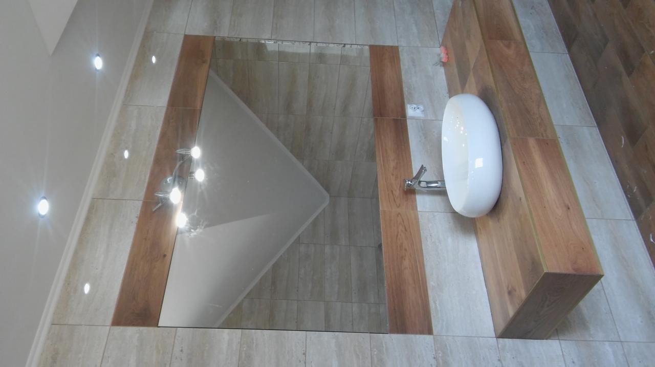 Nowoczesna łazienka z owalną umywalką nablatową na drewnianym blacie, lustro w drewnianej ramie, oświetlenie punktowe w suficie i płytki imitujące kamień.