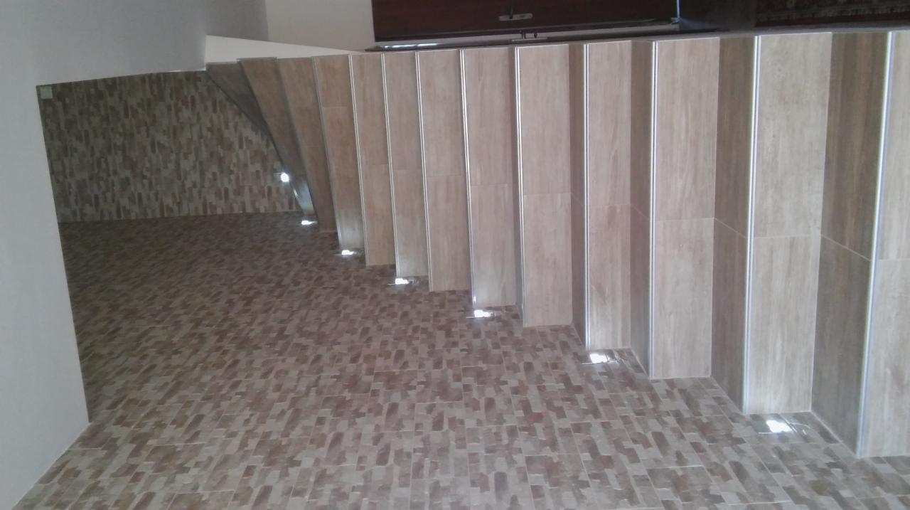 Korytarz z wykończoną podłogą z płytek w odcieniach brązu, z widocznymi pionowymi panelami z jasnego drewna i subtelnym oświetleniem punktowym przy podłodze.