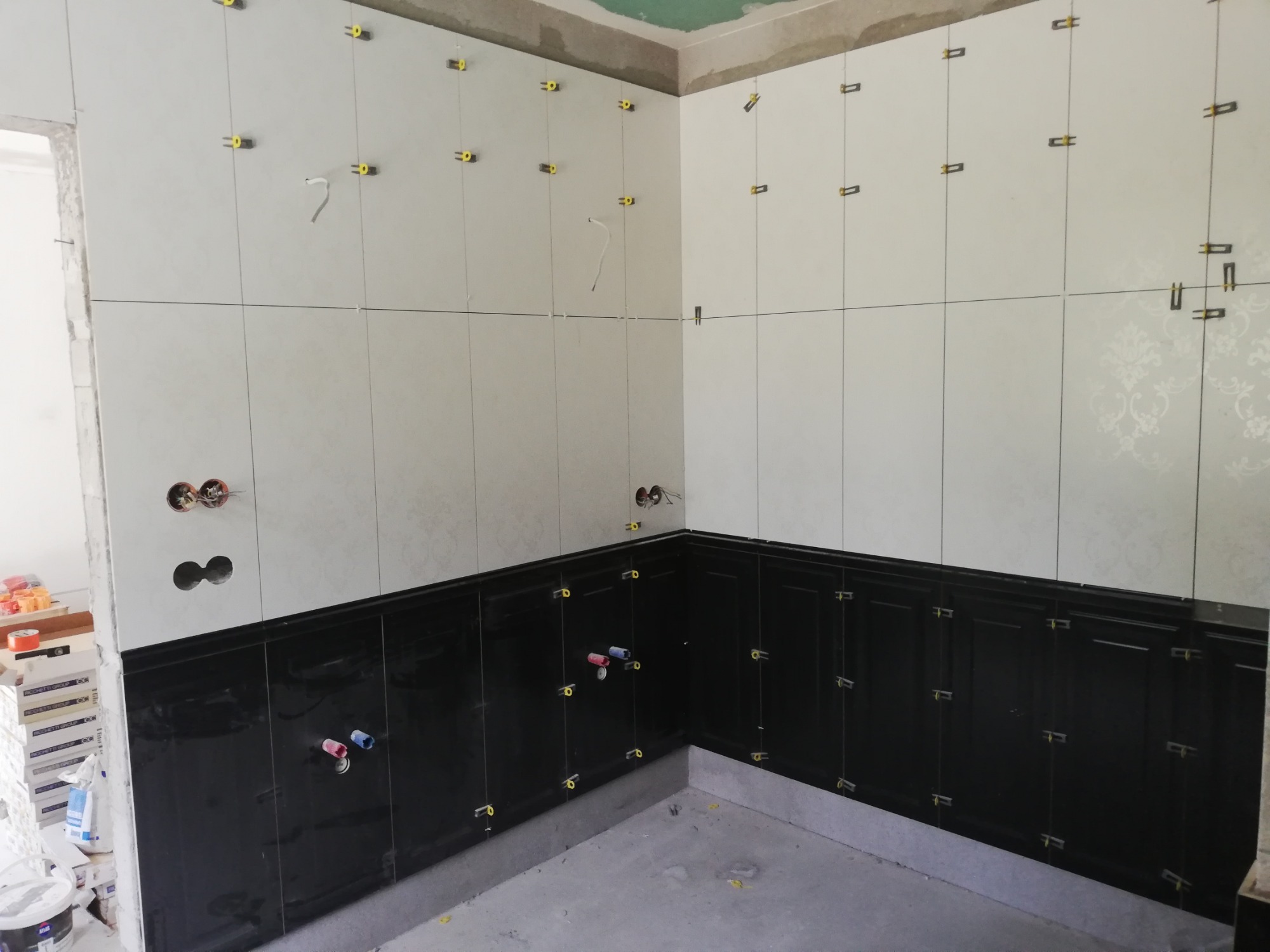 Wnętrze kuchni w trakcie układania czarno-białych płytek ceramicznych na ścianach, z użyciem żółtych krzyżyków dystansowych; widoczne otwory na instalacje.