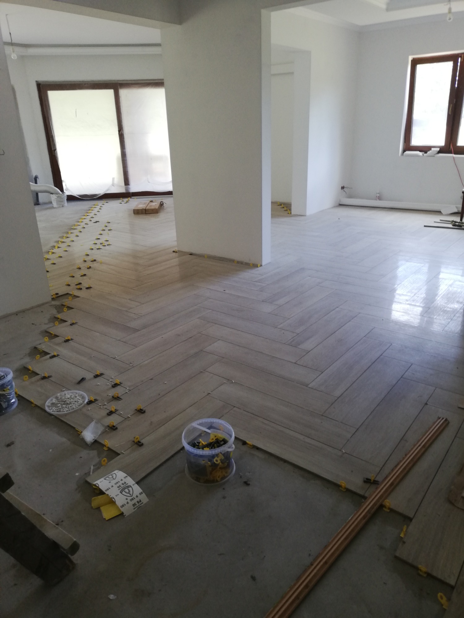 Układanie podłogi z paneli w jodełkę, widoczne plastikowe kliny dystansowe i materiały budowlane, surowy stan wykończenia ścian i sufitu, duże okna.