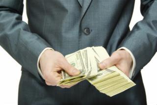 Mężczyzna w szarym garniturze trzyma w dłoniach plik banknotów dolarowych, ukrywając część za plecami, na białym tle.