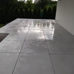 Świeżo ułożona, mokra nawierzchnia z szarych płyt betonowych na tarasie, odbijająca niebo i otoczenie, z widocznym fragmentem drewnianego podestu i rzędem zielonych tuj w tle.