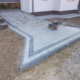 Świeżo ułożona, dwukolorowa kostka brukowa przed garażem nowoczesnego domu, z zagęszczarką w tle, oczekująca na ostateczne wykończenie.