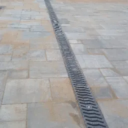 Szara kostka brukowa z liniowym odpływem wody, widoczny betonowy płot w tle.