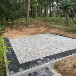 P.H.U. Węglewscy - Ułożona kostka brukowa w leśnej scenerii. Jasnoszara kostka otoczona ciemniejszym pasem, tworząca plac z niskimi schodkami. Widoczne drzewa i naturalne otoczenie.