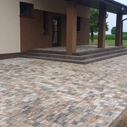 P.H.U. Węglewscy - Nowoczesny taras z kostki brukowej w odcieniach beżu i brązu, prowadzący do wejścia domu z kolumnami. Widoczne schody z kostki i fragment ogrodu w tle.