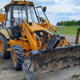 P.H.U. Węglewscy - Żółta koparko-ładowarka JCB 3CX na placu budowy, widoczna sterta ziemi i budynek w trakcie budowy, błękitne niebo z chmurami w tle.
