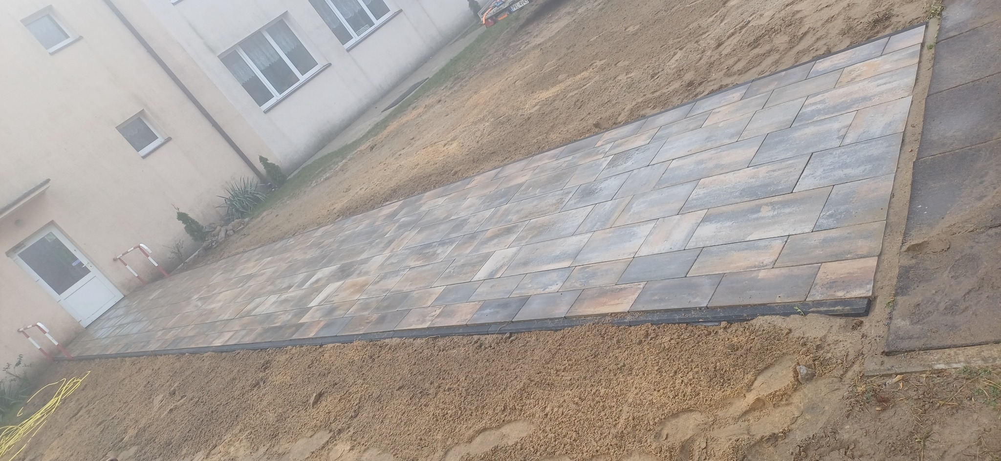 Świeżo ułożona, prostokątna ścieżka z kostki brukowej w odcieniach szarości i brązu, otoczona piaskiem, prowadząca wzdłuż jasnej ściany budynku z białymi oknami i drzwiami.