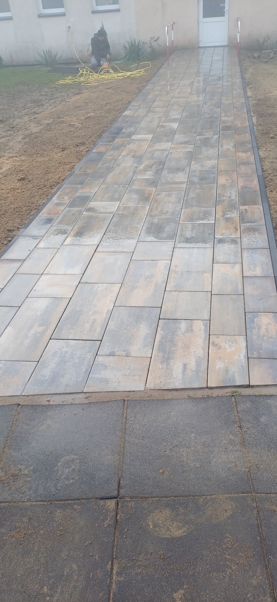 Świeżo ułożona ścieżka z kostki brukowej prowadząca do budynku, z pracownikiem wygładzającym powierzchnię w tle oraz zwiniętym żółtym kablem obok.