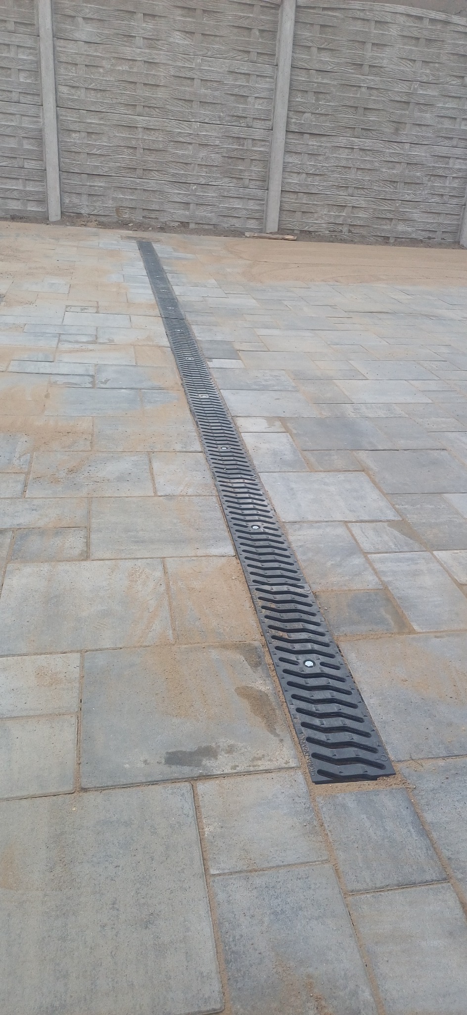 Ułożona szara kostka brukowa z długim, czarnym odpływem liniowym pośrodku, na tle betonowego ogrodzenia.