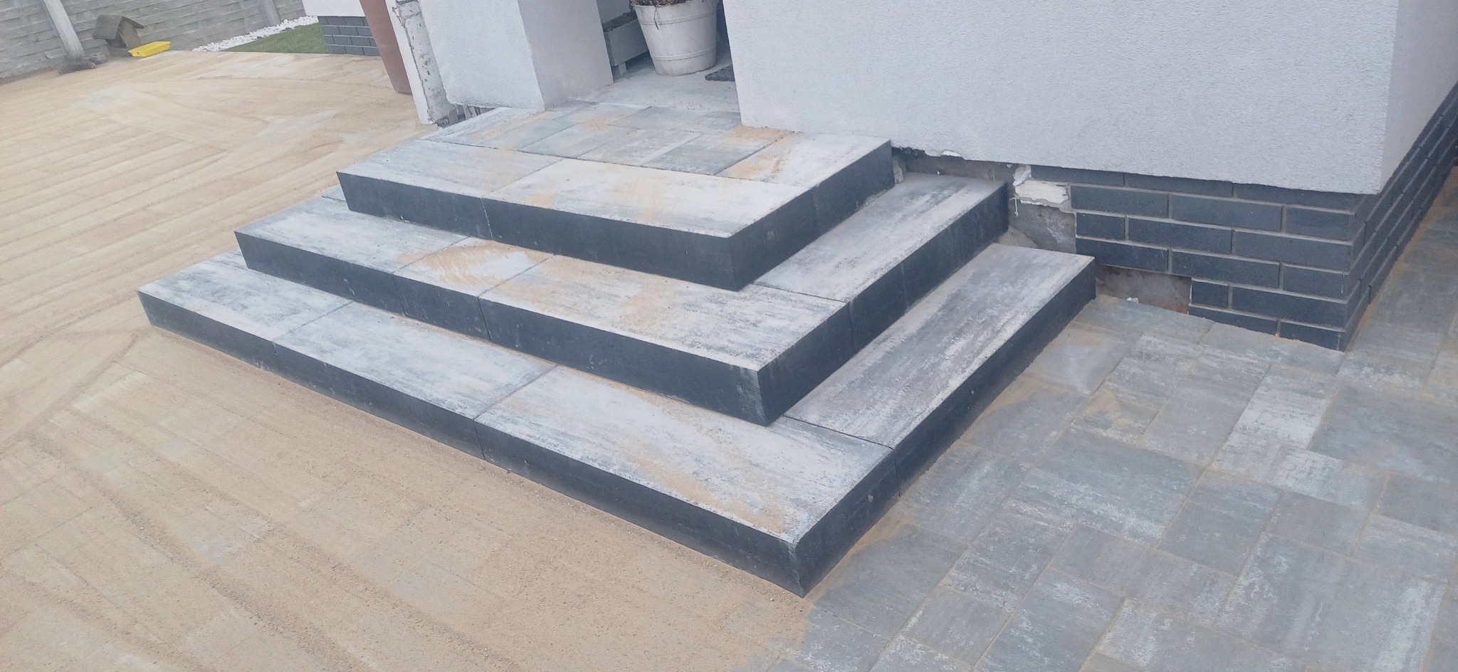 Trzy stopnie schodów wejściowych wyłożone szarą kostką brukową, otoczone piaskiem i szarą kostką, z fragmentem ściany budynku w tle.