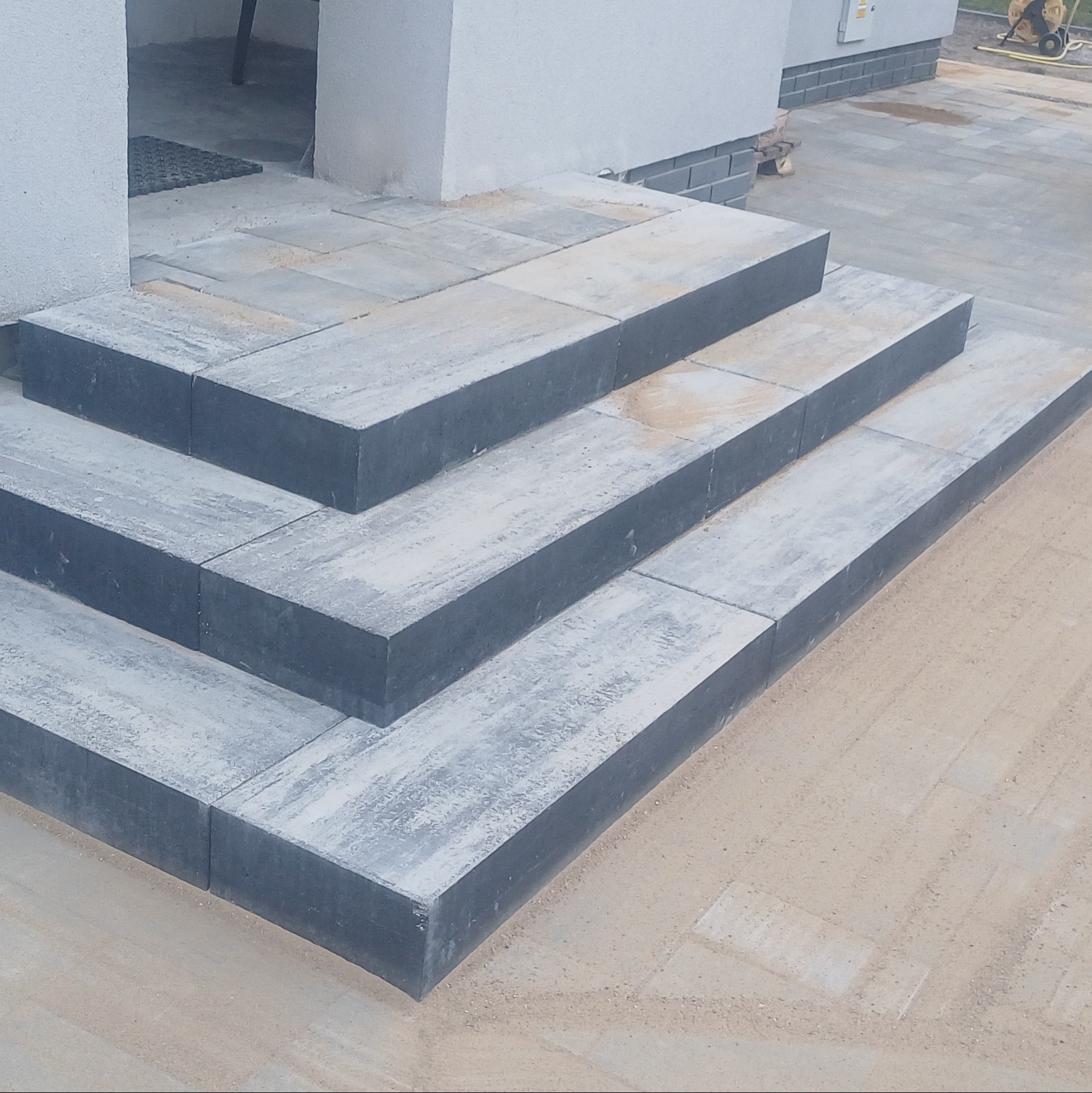 Schody wejściowe ułożone z szarej kostki brukowej, prowadzące do budynku z jasną elewacją i fragmentem chodnika z kostki. Widoczne fugi wypełnione piaskiem.