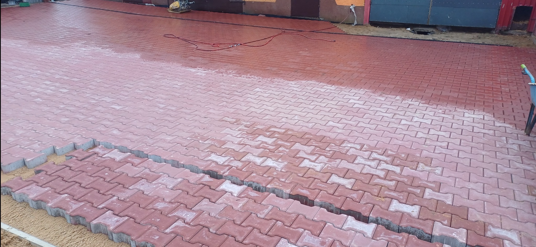 Układanie czerwonej kostki brukowej typu 'I', widoczny postęp prac, część powierzchni wyłożona, część jeszcze w trakcie, piasek pod kostką, lekko mokra powierzchnia kostki, w tle widoczny...