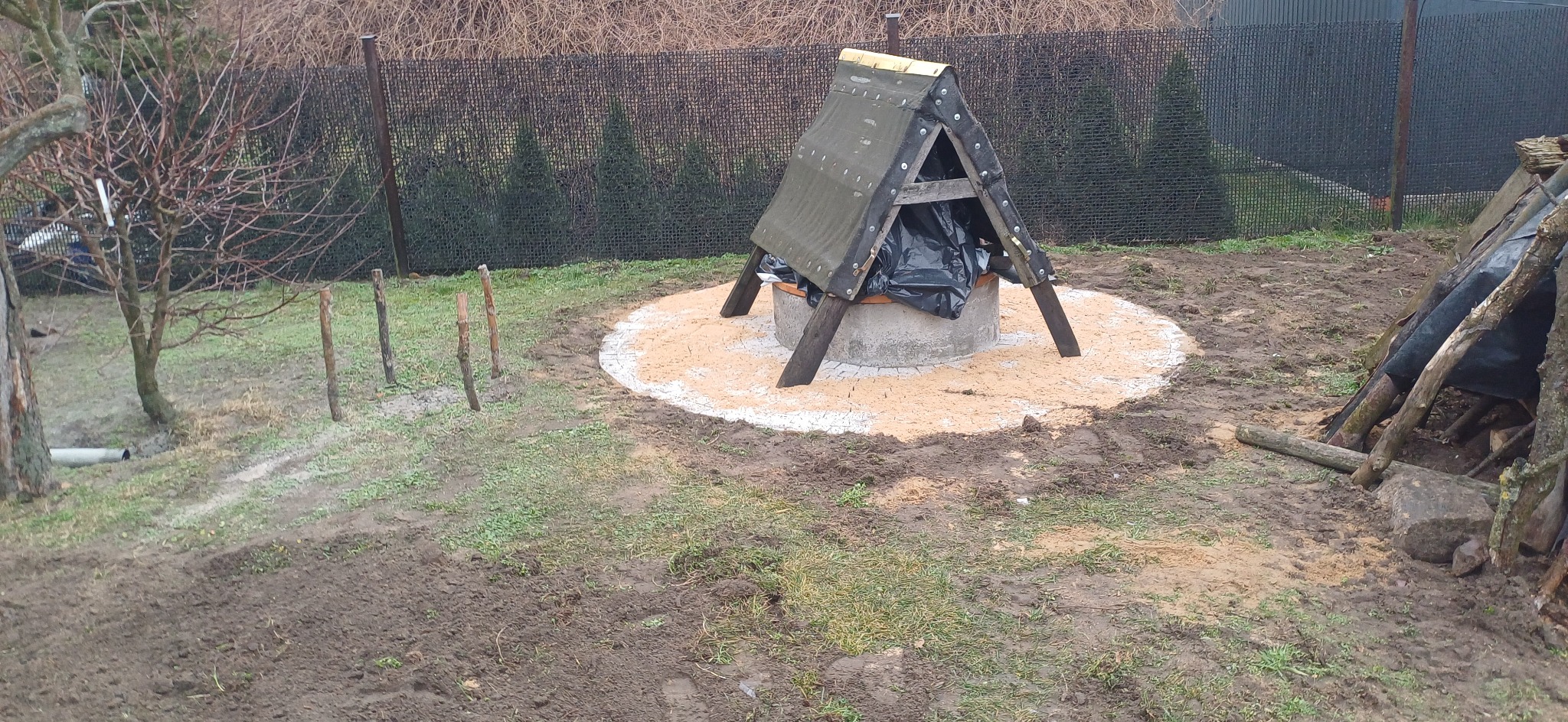 Betonowa studnia z drewnianą konstrukcją ochronną, otoczona piaskiem, na nierównym, błotnistym terenie.