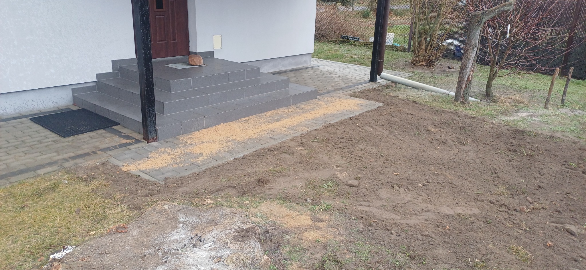 Ułożona kostka brukowa przed domem, widoczny fragment wyrównanego terenu z przygotowaniem pod dalsze prace. Na schodach leży kamień.