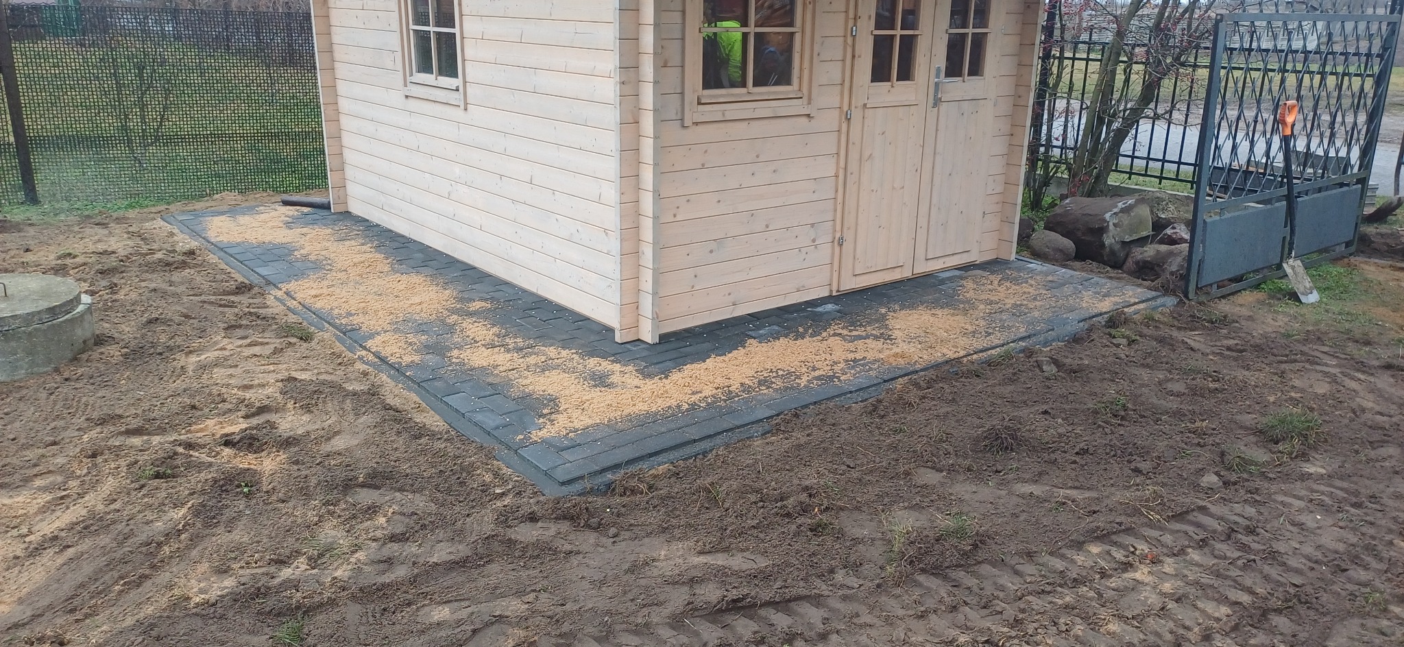 Ułożona szara kostka brukowa przed drewnianą altaną, posypana trocinami, widoczny teren z ziemią i fragment ogrodzenia.