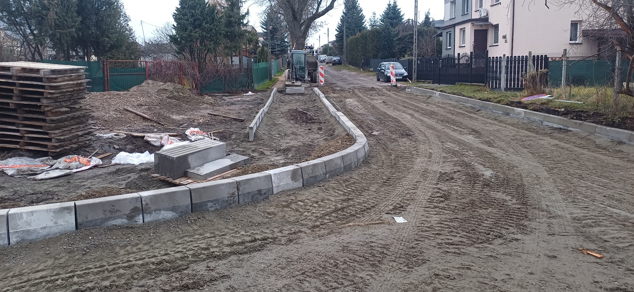 Przygotowanie podłoża pod chodnik z obrzeżami betonowymi, widoczna mini koparka, palety z materiałami i ślady opon na piasku.