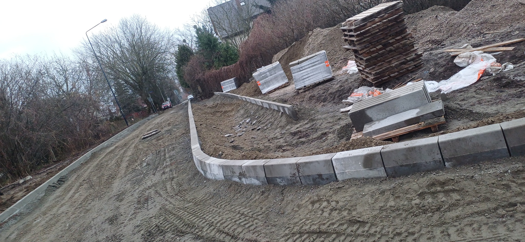 Krawężniki betonowe ułożone wzdłuż krawędzi drogi gruntowej, palety z kostką brukową w tle, widoczne ślady opon.