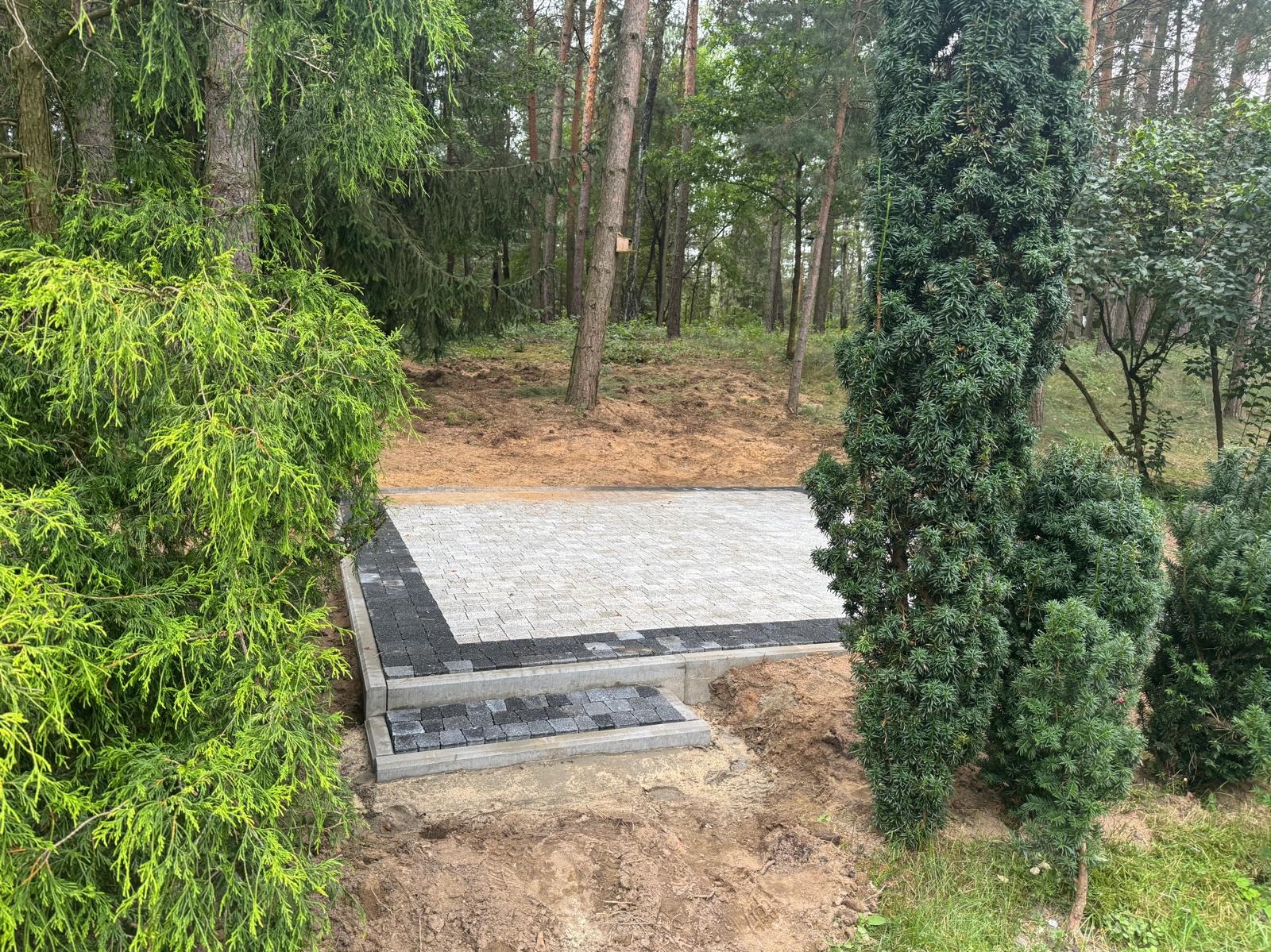 Ułożona kostka brukowa w leśnym otoczeniu, z obrzeżami z ciemnej kostki i schodami. Widoczne drzewa i krzewy, świeżo wykończona nawierzchnia.