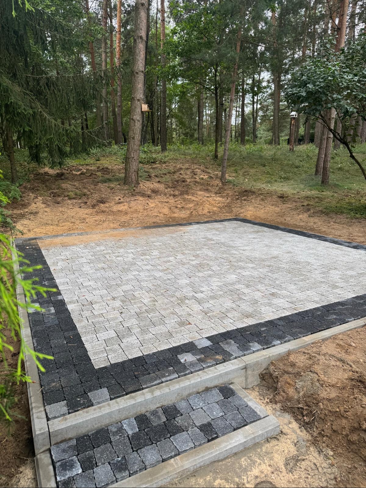 Ułożona kostka brukowa w leśnej scenerii. Jasnoszara kostka otoczona ciemniejszym pasem, tworząca plac z niskimi schodkami. Widoczne drzewa i naturalne otoczenie.