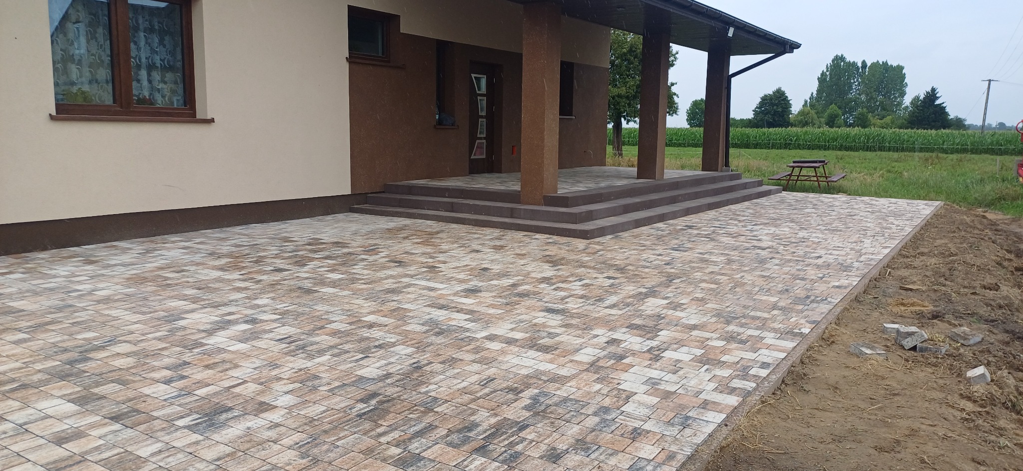 Nowoczesny taras z kostki brukowej w odcieniach beżu i brązu, prowadzący do wejścia domu z kolumnami. Widoczne schody z kostki i fragment ogrodu w tle.