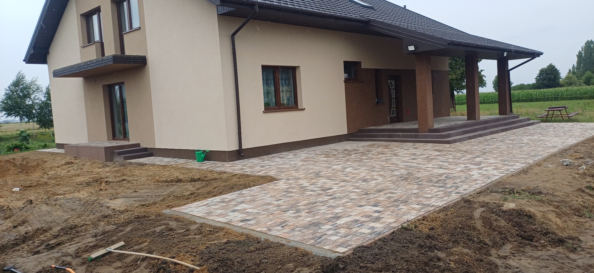 Nowa, beżowo-brązowa kostka brukowa przed domem z zielonym akcentem konewki. Widoczny fragment ogrodu i przygotowany teren pod dalsze prace.