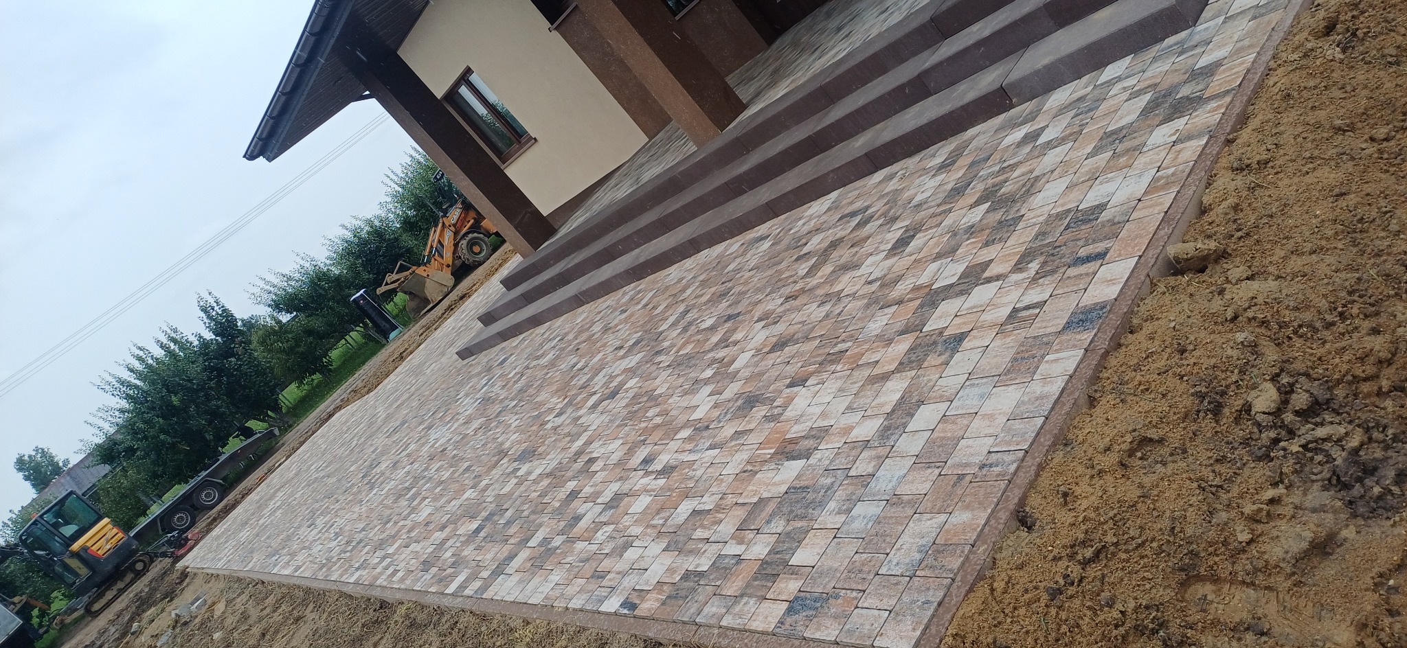 Ukończony podjazd z kostki brukowej w odcieniach beżu i brązu, prowadzący do schodów wejściowych domu. W tle widoczny sprzęt budowlany i zieleń.