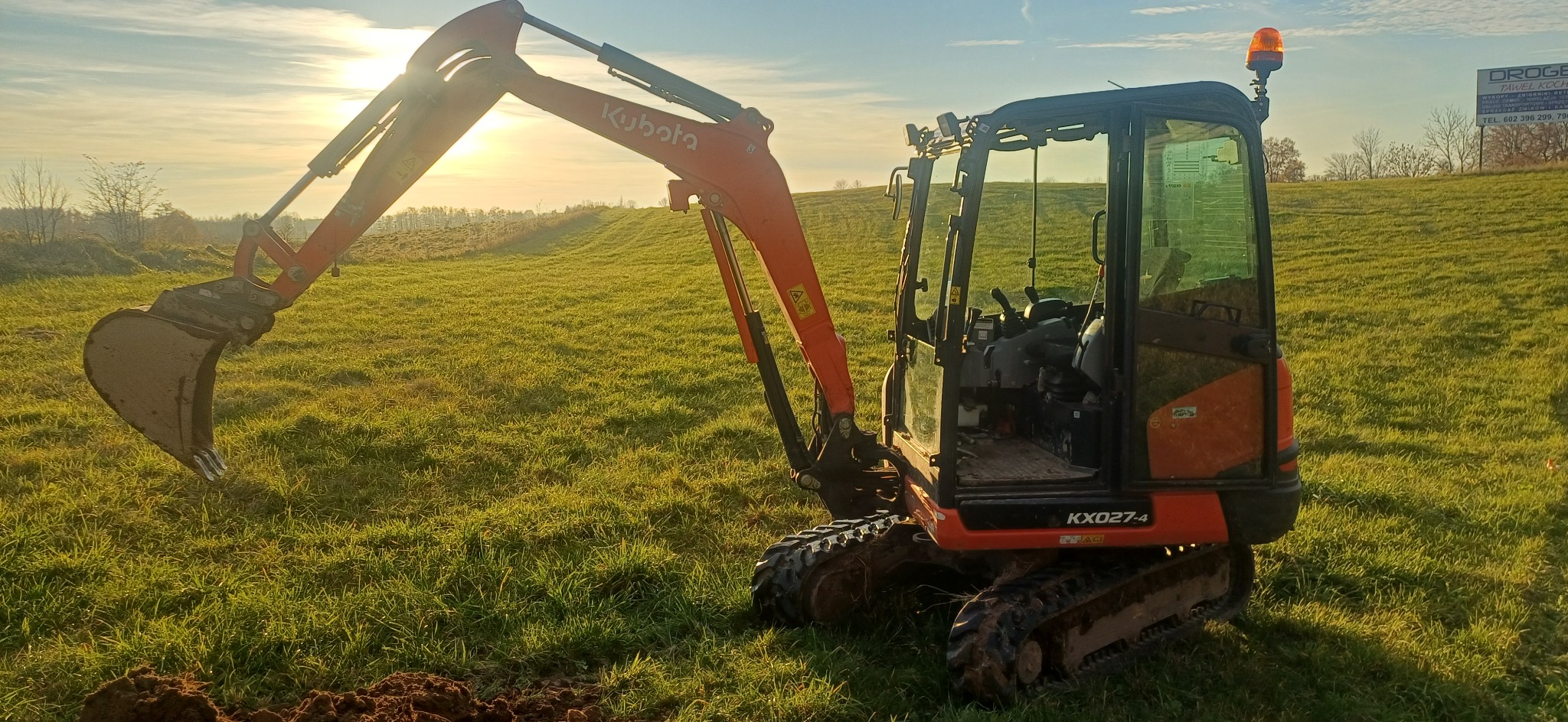 Pomarańczowa mini koparka Kubota KX027-4 na zielonej łące w promieniach zachodzącego słońca. Widoczny wykop ziemi i tablica reklamowa w tle.