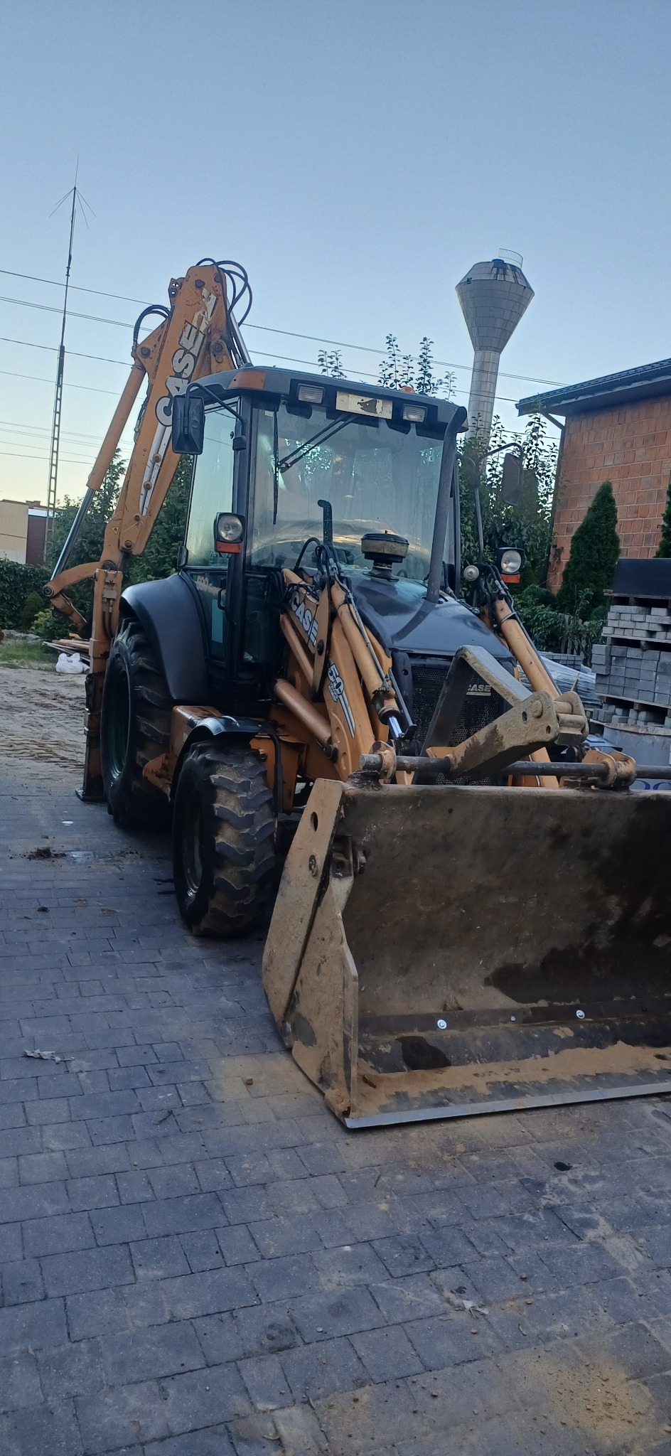 Żółto-czarna koparko-ładowarka Case na kostce brukowej, z widoczną wieżą ciśnień w tle. Sprzęt budowlany po pracy, z zabrudzeniami na łyżce i oponach.